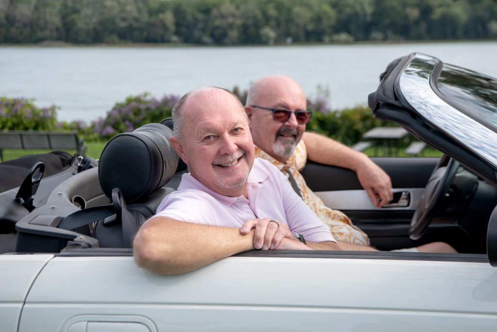 photo shows two men around age 65, smiling at the camera and sitting in a convertible with the top down. It’s a sunny day, and they are parked near a lake with greenery all around
